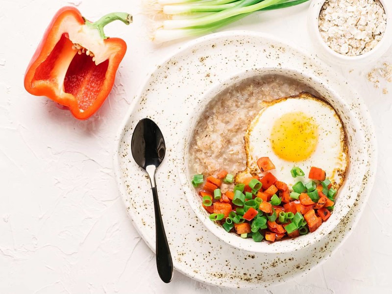 Savory oatmeal in craft trendy bowl, served with vegetables and fried egg on white concrete background. Red bell pepper and bunch onion on background. Top view or flat lay. Copy space