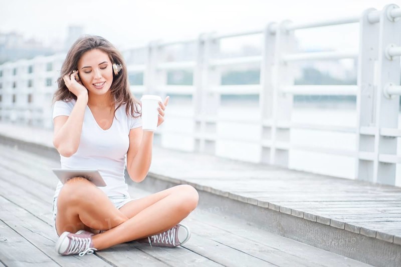 Happy young woman with music headphones and a cup of coffee to go, surfing the internet on a tablet PC, listening to music and sitting on the bridge