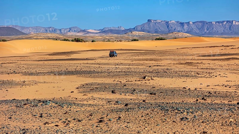 An off-road vehicle moves through a vast landscape of sand dunes and mountains, With off-road vehicles through the endless landscape of the Sahara desert in Algeria