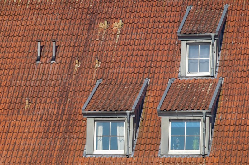 Close-up view of a sloped roof featuring red clay tiles, three dormer windows, and two chimneys