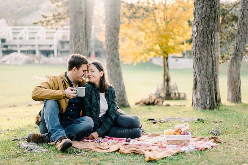 Husband and wife sit on a bedspread with their foreheads leaning next to a basket of food.
