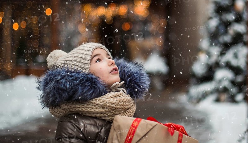 Happy girl with a gift box for Christmas catches snowflakes with his tongue on a city in winter with snow on festive market with decorations and lights. Warm clothes, knitted hat, scarf and fur