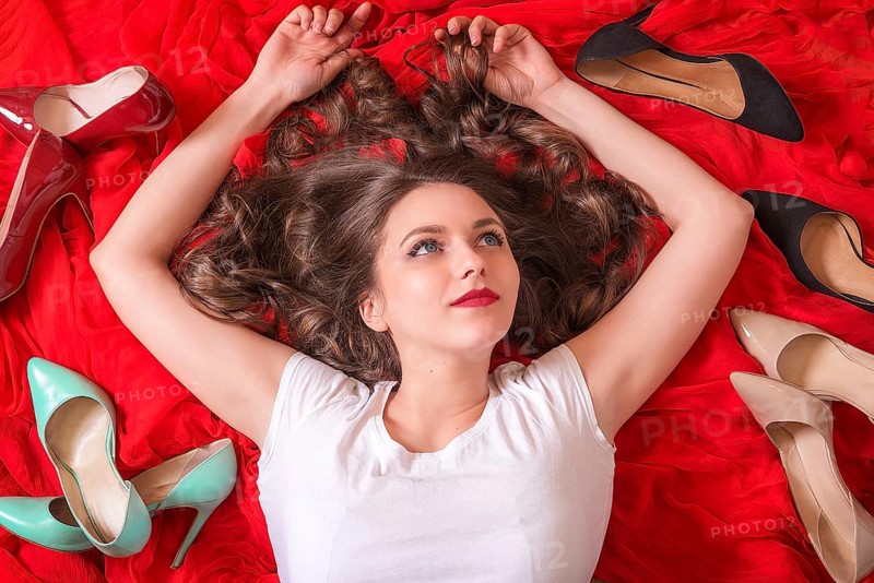 Beautiful woman with blue eyes and red lips, lays on a red fabric, smiling and surrounded by many elegant shoes in a variety of colors