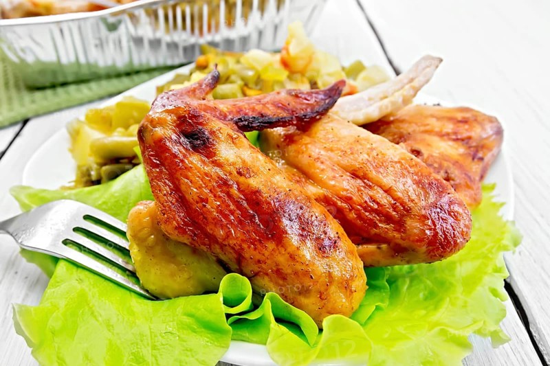 Fried chicken wings with vegetables and salad leaves on a plate, fork on the background light wooden boards