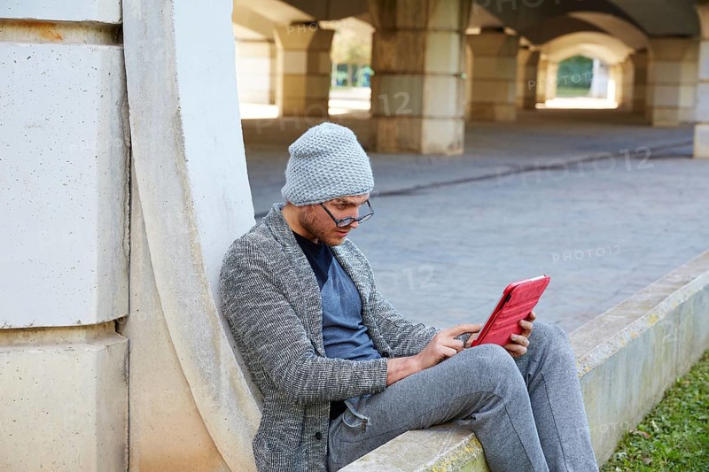 Modern young man with tablet pc touch working under a bridge with wool winter cap