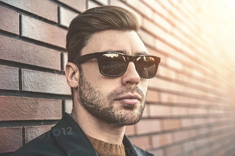 Portrait of stylish handsome young man with bristle standing outdoors and leaning on brick wall. Man wearing jacket, sweater and sunglasses