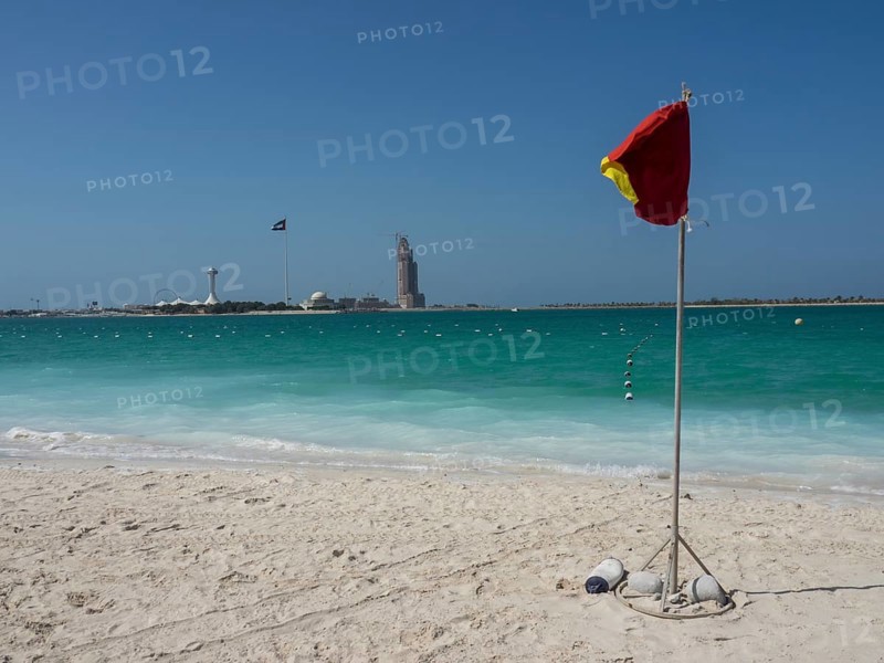 A red warning sign stands on the beach with turquoise water and a blue sky in the background, shimmering green sea with beach, parasols and sunbeds in front of a bright blue sky, Abu dhabi, Arab Emirates