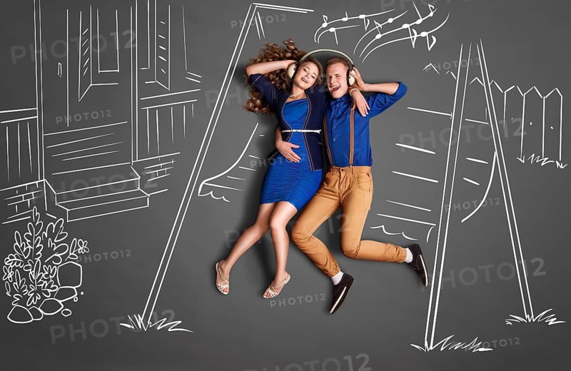 Happy valentines love story concept of a romantic couple sitting on the canopy swing sharing headphones and listening to the music against chalk drawings backyard background