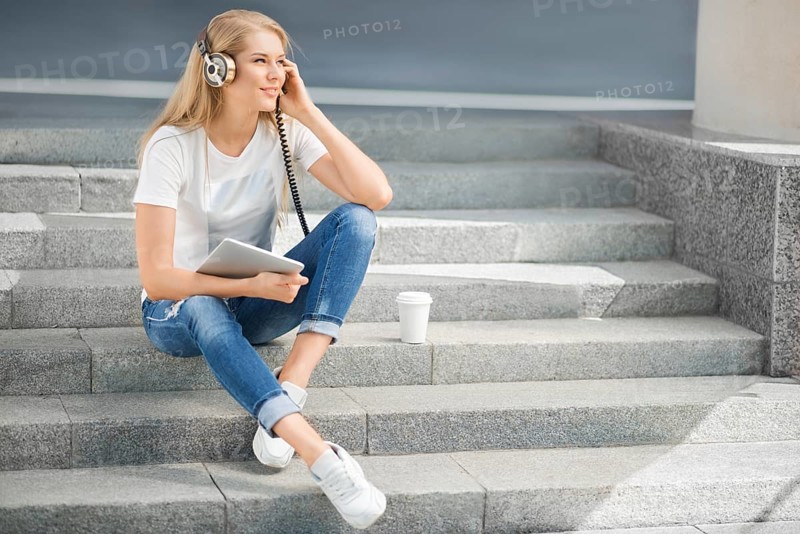 Happy young woman with vintage headphones and a cup of coffee to go, surfing the internet on a tablet PC, listening to music and sitting on a staircase in front of an urban background