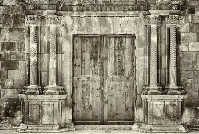 Monochrome sepia ancient wooden double doors in an old stone building with crumbling ornate columns surrounding the entrance