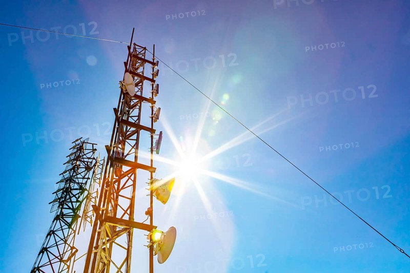 A low angle view of two mobile communication data transmitter and GPS signal towers, against a blue sky with bright sun shine and colorful lens flare