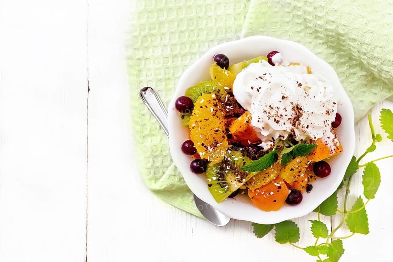 Fruit salad with orange, kiwi, cranberries and baked pumpkin, whipped cream, sprinkled with chocolate and coconut with mint in a bowl on a towel on wooden board background from above