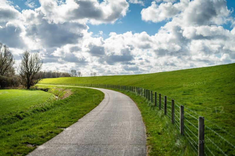An empty country lane winds along green meadows and a fence under a cloudy sky, cycle path, Leer, East Frisia, Lower Saxony