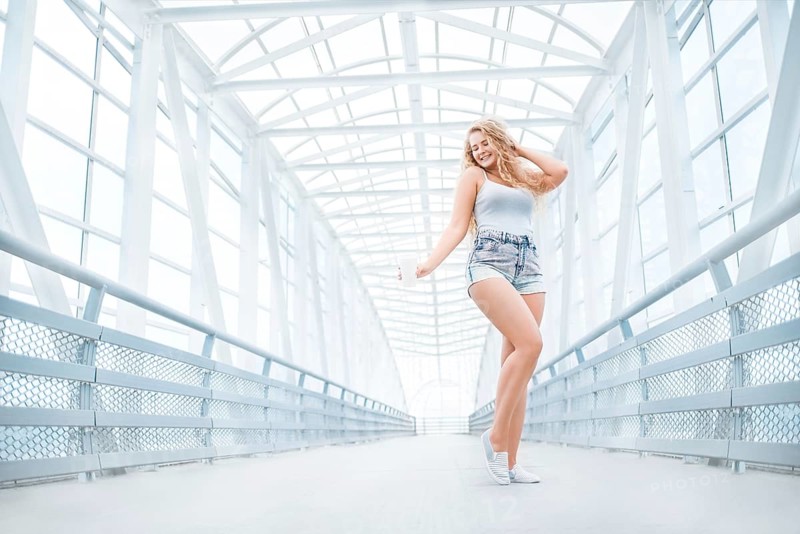 Beautiful young woman with long curly hair holding a takeaway cup of coffee and standing on the bridge against an urban background