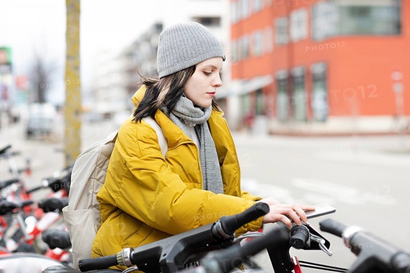 Woman in yellow jacket and cap on rental bikes in city street, cold winter day, environmentally friendly