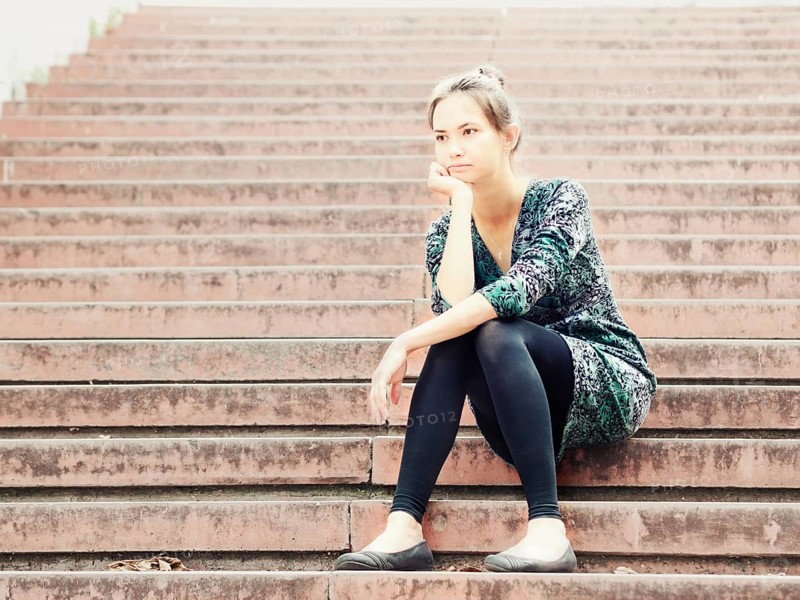 Sad lonely young woman sitting on steps. Portrait of serious girl with facial expression outdoors