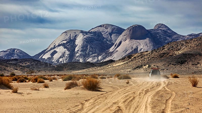 A vehicle moves along a dusty track through a vast desert landscape, travelling through the Sahara in Algeria by off-road vehicle