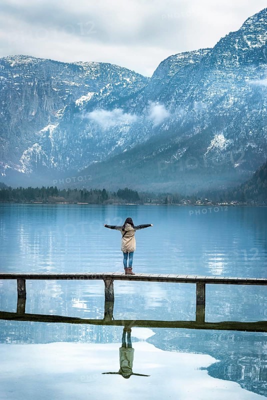 Girl with her arms open, standing on a narrow wooden deck, enjoying the view of the Austrian Alps mountains reflected in the Hallstatter lake water, in Austria