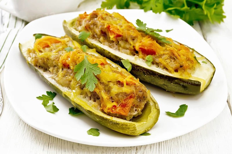 Cucumbers stuffed with minced meat, tomatoes, pepper with cheese and parsley in a plate, cream and greens sauce in a gravy boat, towel on the background of light wooden board