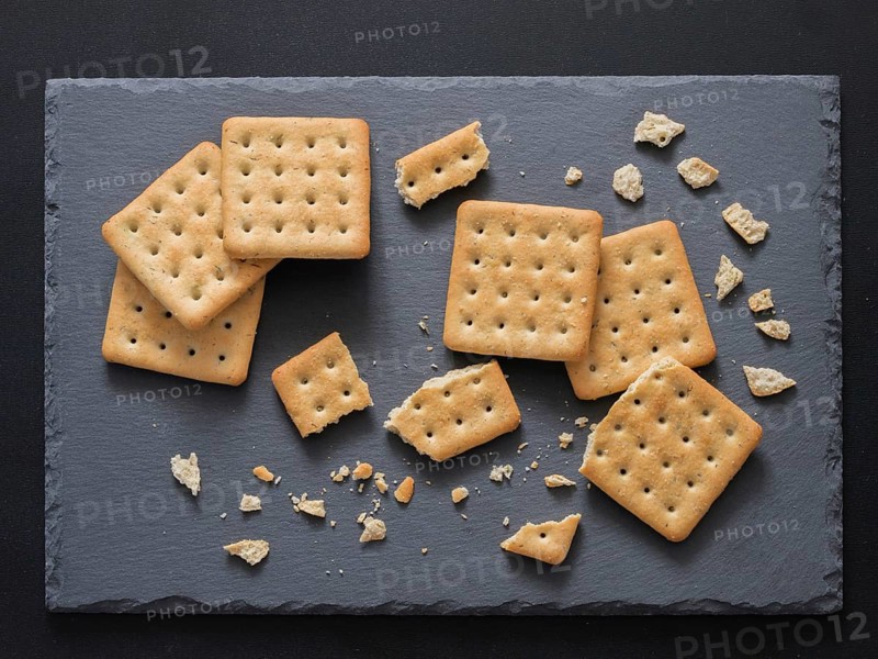 Square crackers with pieces and crumbs on slate gray background. Dry salt cracker cookies with fiber and dry spices. Top view or flat lay
