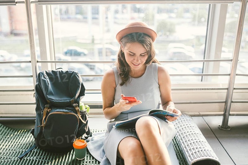 Theme of tourism and travel of young student. Beautiful young caucasian girl in dress and hat sits on floor tourist rug inside terminal airport terminal. Waiting room delayed flight, delay departure