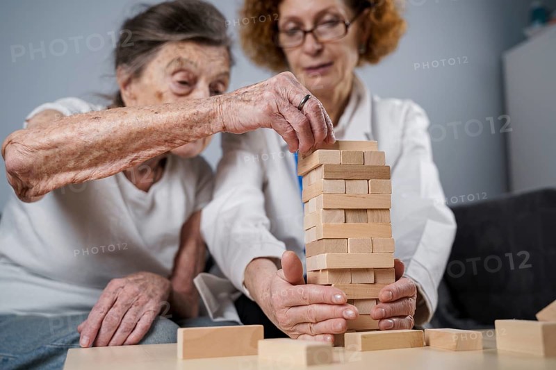 Senior woman practice skills, build wooden blocks, build tower and try not to let it fall, Jenga game. Old patient pull out block, place on top, support doctor during therapy dementia in house