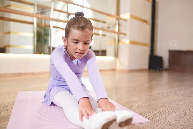 Adorable photo of little ballerina in a lilac dress and white tights stretching exercises in a hall with large mirrors