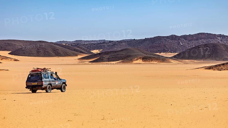 An off-road vehicle stands in vast sand dunes under a clear, sunny sky, off-road through the landscapes of the Sahara in Algeria