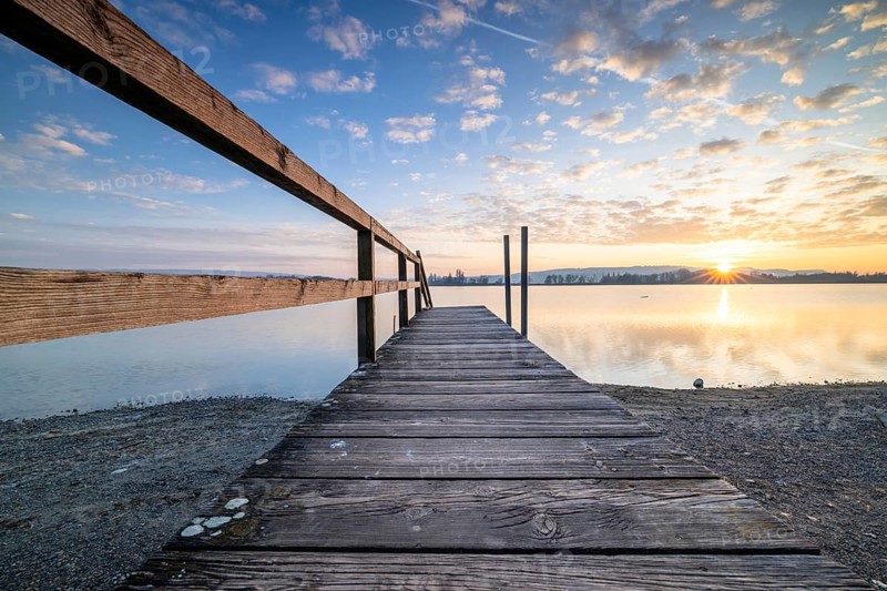 A wooden footbridge leading onto a calm lake as the sun sets under a cloudy sky, Naturfreundehaus, Markelfingen, Untersee, Lake Constance, Baden-Württemberg, Germany, Europe
