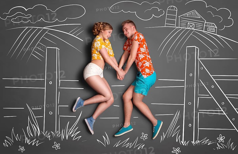 Happy valentines love story concept of a shy romantic couple sitting on a hedge and holding hands against chalk drawings background of a landscape in summer