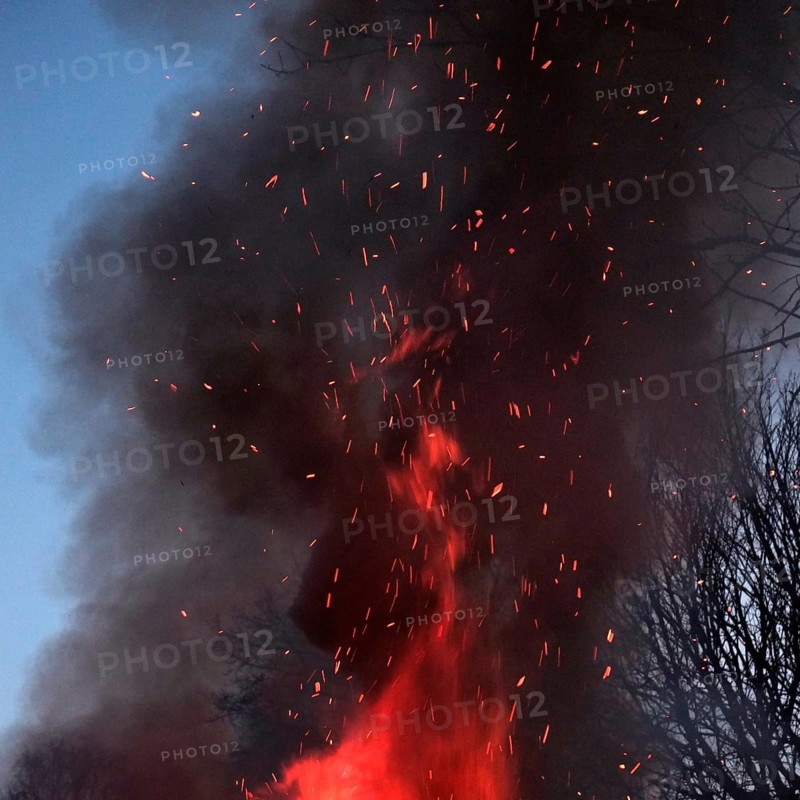 Crackling flames eat into the high pile of an Easter fire, dark smoke with flying sparks rises and darkens the surroundings. Square format. Crackling flames move through a high-piled heap of an Easter Saturday bonfire upwards, dark smoke with flying sparks rises up and obscures the environment. Square Format