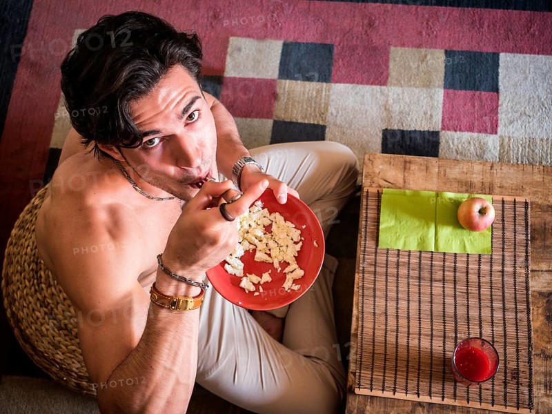 From above shot of happy muscular shirtless man sitting on floor having breakfast eating cereal, smiling at camera