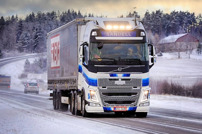 Blue white Volvo FH semi trailer truck Kuljetusliike Harri Sandell Oy with bright headlights on road 52 at winter dusk. Salo, Finland. Dec 27, 2021