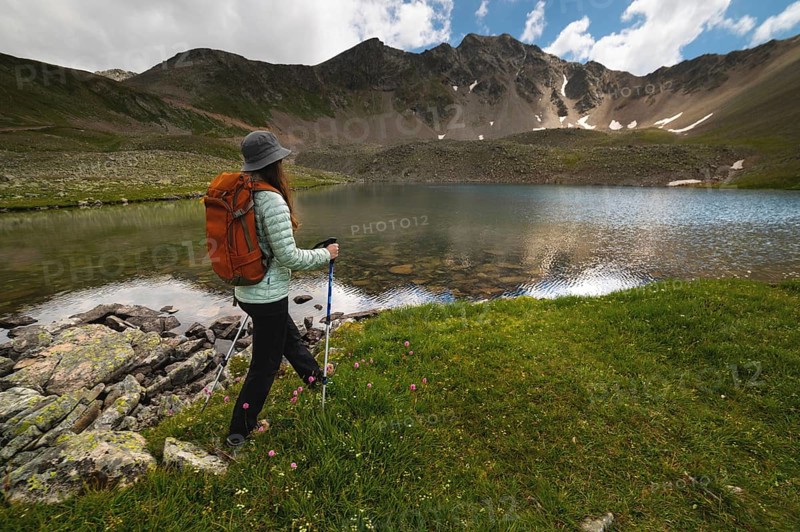A woman with an orange backpack and a hat goes to a transparent lake in the mountains on a clear day