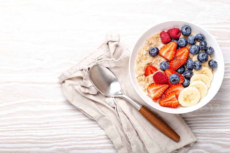 Oatmeal porridge with fruit and berries in bowl with spoon on white wooden background table top view, homemade healthy breakfast cereal with strawberry, banana, blueberry, raspberry. Space for text, food photography, food photography