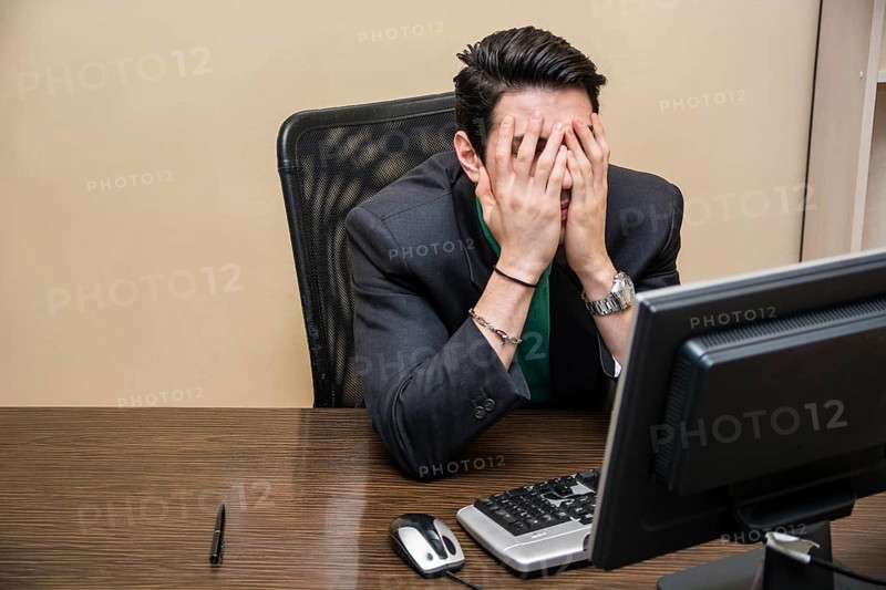 Desperate, worried young businessman sitting at his desk in front of his computer with his face in his hands and eyes closed