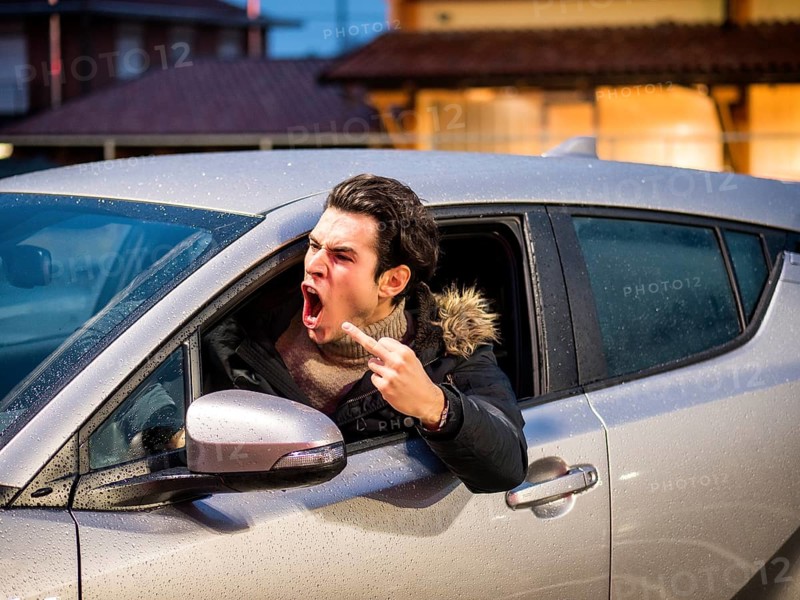 Aggressive man driving car and screaming away while showing obscene gesture on street