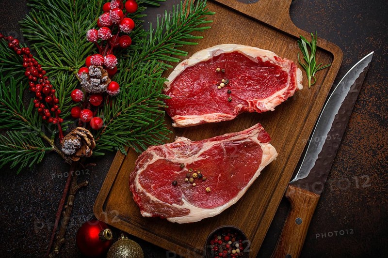Two raw uncooked meat beef steaks on wooden cutting board with knife and seasonings on dark rustic background with Christmas festive decoration from above, food photography, food photography
