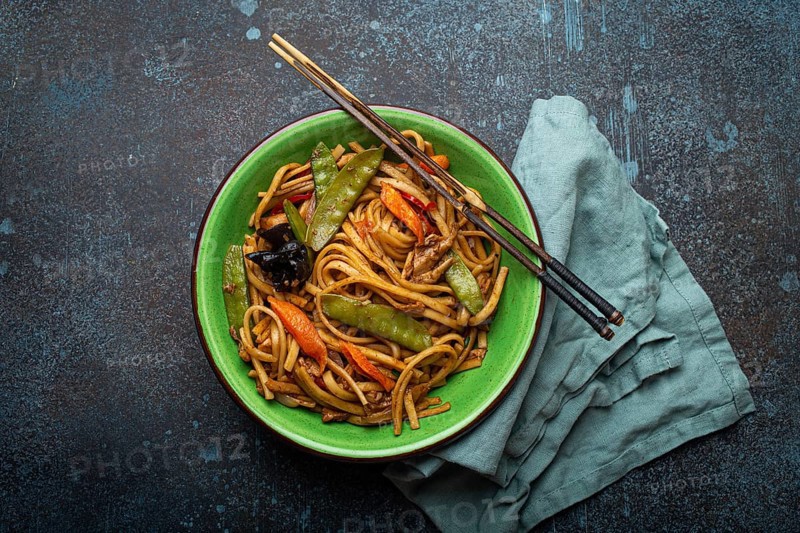 Asian dish stir fry udon noodles with chicken, vegetables and mushrooms in green ceramic bowl with wooden chopsticks on rustic dark blue concrete background from above, Chinese or Thai fast food, food photography