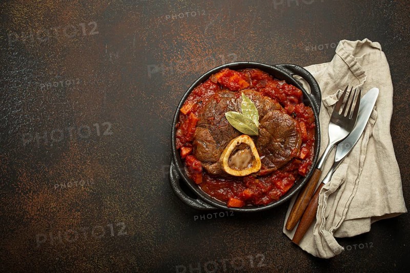 Traditional Italian dish Ossobuco all Milanese made with cut veal shank meat with vegetable tomato sauce served in black casserole pan top view on rustic brown background, copy space, food photography, food photography
