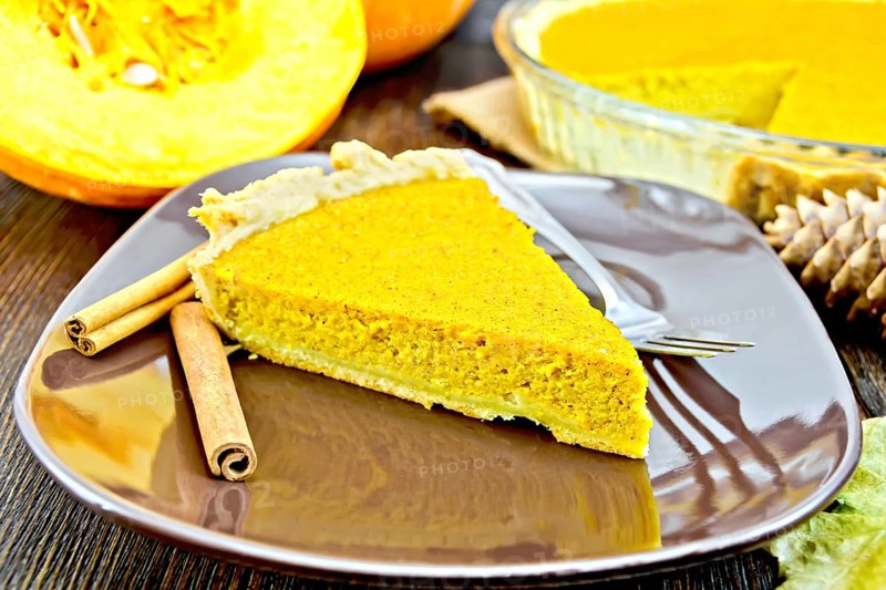 A piece of pumpkin pie in a brown plate with a fork and cinnamon, pumpkin, cones and maple leaf on the background of dark wood planks