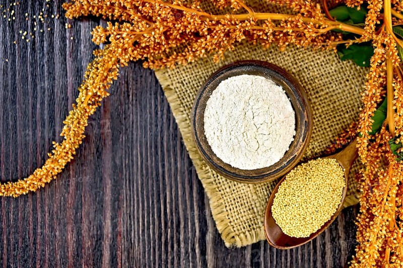 Flour amaranth in a clay bowl, a spoon with grain, brown flower with leaves on a napkin from a sacking on a background of wooden boards on top