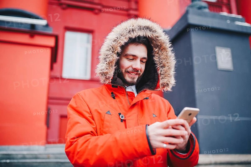 Handsome young male student with toothy smile and beard stands on red wall background, facade of educational institution in red winter jacket with hood with fur, Uses finger on screen of mobile phone