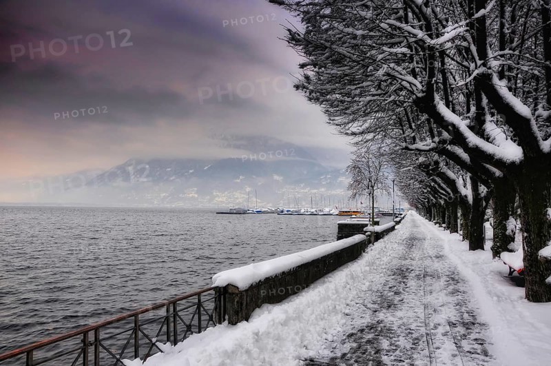 Walkway and Bare Trees with Snow in Winter on the Waterfront to Lake Maggiore with Mountain and Beautiful Storm Clouds in Locarno, Ticino, Switzerland, Europe