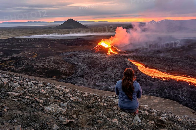 A woman sits on a mountain and looks at a glowing volcano, Grindavik, Sudurnes, Iceland, Europe