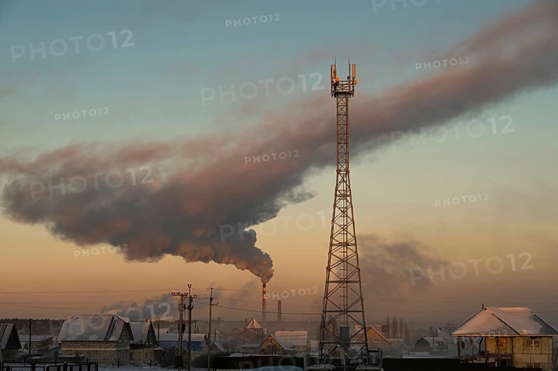 Smoke rises from the chimney of the boiler room over the city and the cell tower is in the foreground. Russia, Novosibirsk region, air temperature -41 degrees Celsius