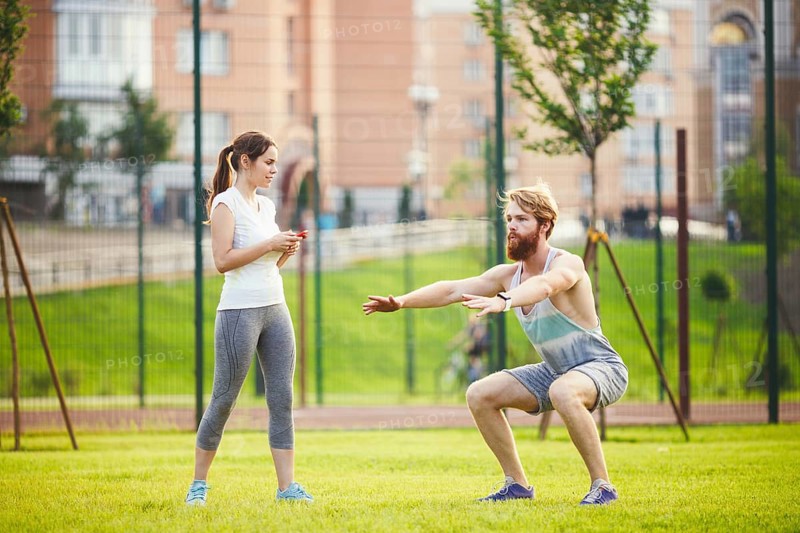 Team work in sports. female trainer training man with beard in park on green grass, lawn at sunset. The girl holds phone in hand and uses stopwatch, guy does exercises for the buttocks of sitting down