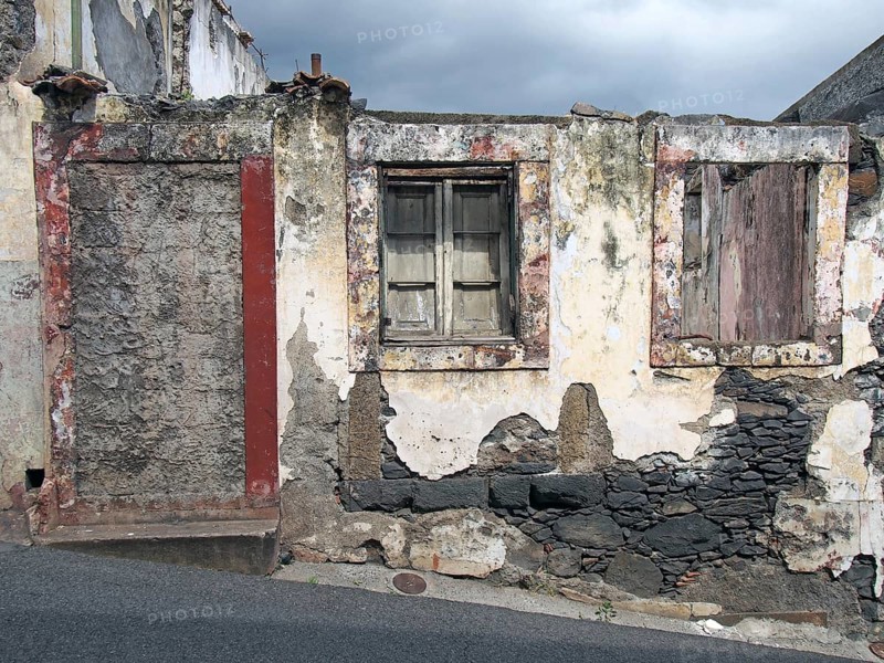 An old ruined house partly collapsed on a sloping street with a blocked up door crumbling walls and fading red painted windows with closed shutters