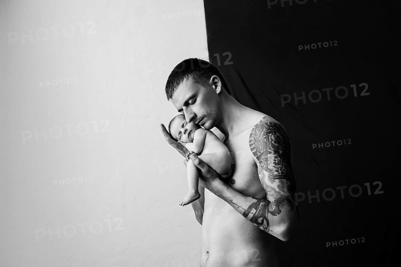 Father with tattoo on his arm is holding his newborn baby on a black and white background.