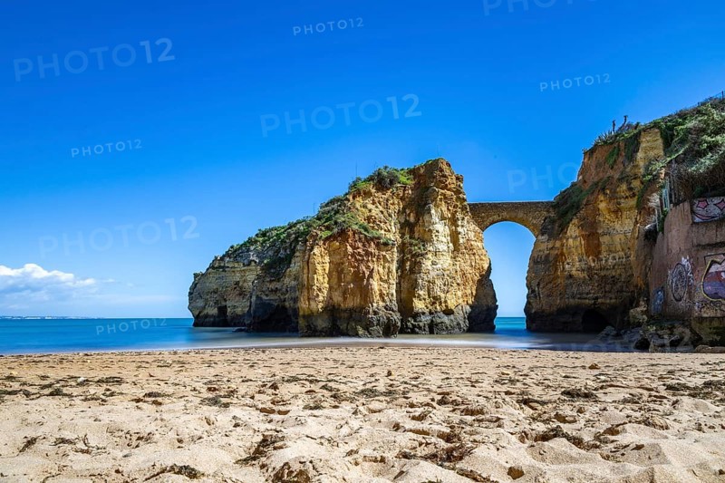 Yellow rocks and cliffs on a sandy beach. A bay by the sea, over which the old stone bridge of the Pinhão fortress connects two cliffs. Praia da Caldeira, Lagos. Algarve, Portugal, Europe